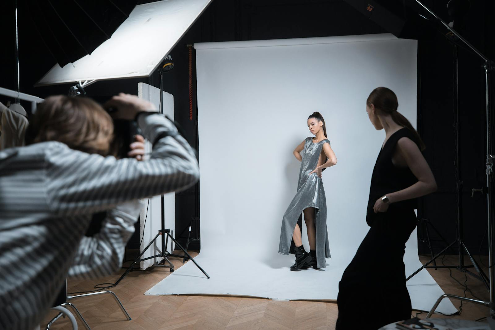 A working set in the Lumina Studio — two stylists pinning a garment on a model while a photographer reviews tethered captures on a calibrated monitor.